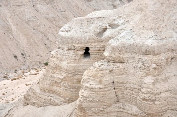 Cave 4 at Qumran: a dark opening high in a pale, layered rock cliff above a dry valley.